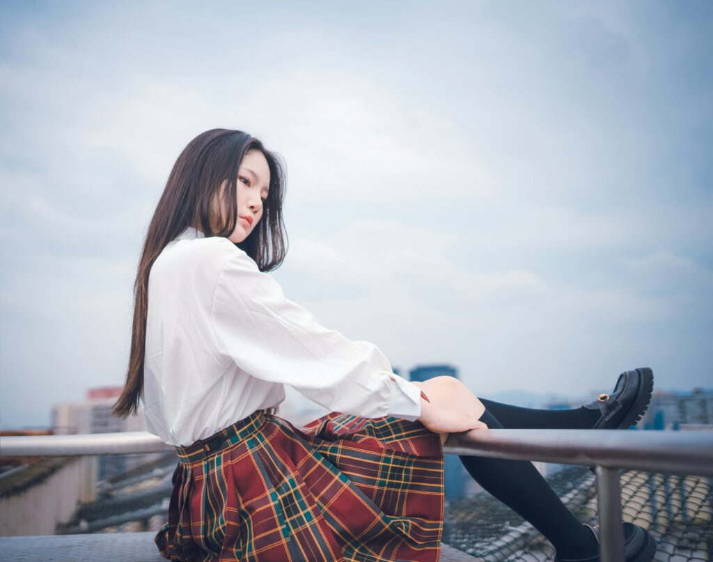 A young woman in a white, long-sleeved shirt and an orange and green plaid skirt sits on a railing.