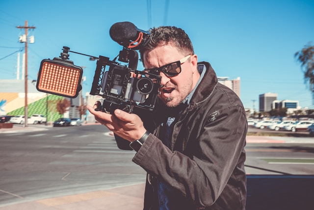 A man holds a video camera in an empty street. 
