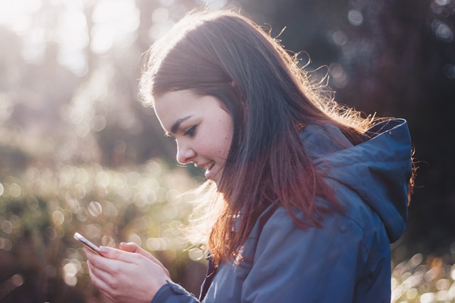 A young teen stands outdoors and smiles as she browses TikTok on her phone.