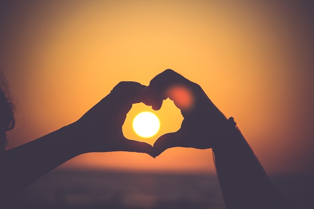Due mani formano un cuore intorno al sole che tramonta. 