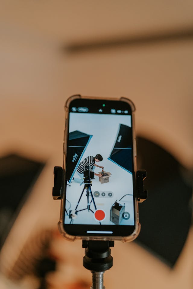 A cell phone on a tripod is recording a man who is unboxing a package.