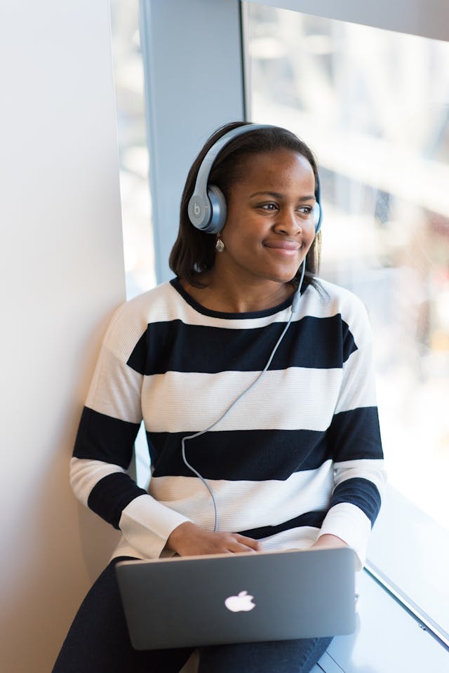A woman in a striped shirt listens to headphones connected to her laptop.
