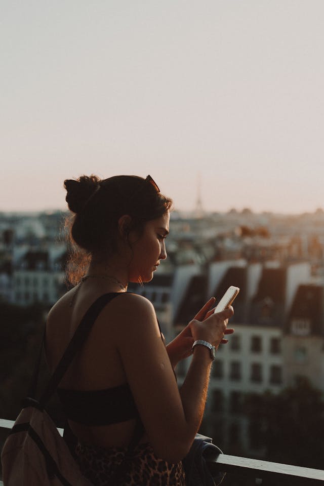 A woman stands on a balcony and scrolls through her cell phone.
