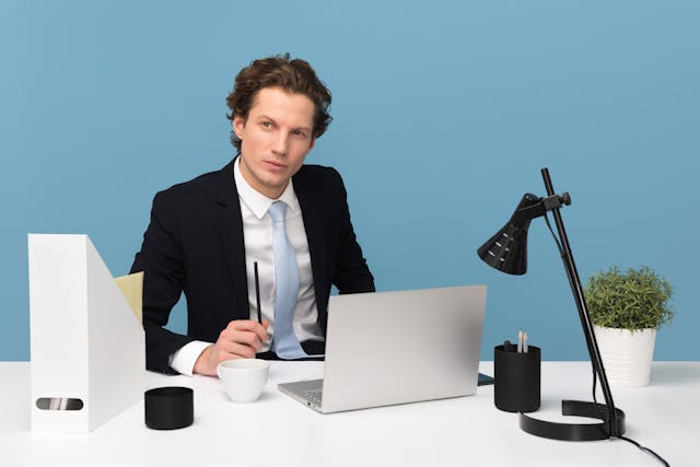 A man in a suit and tie sits in front of his laptop and stares into space.