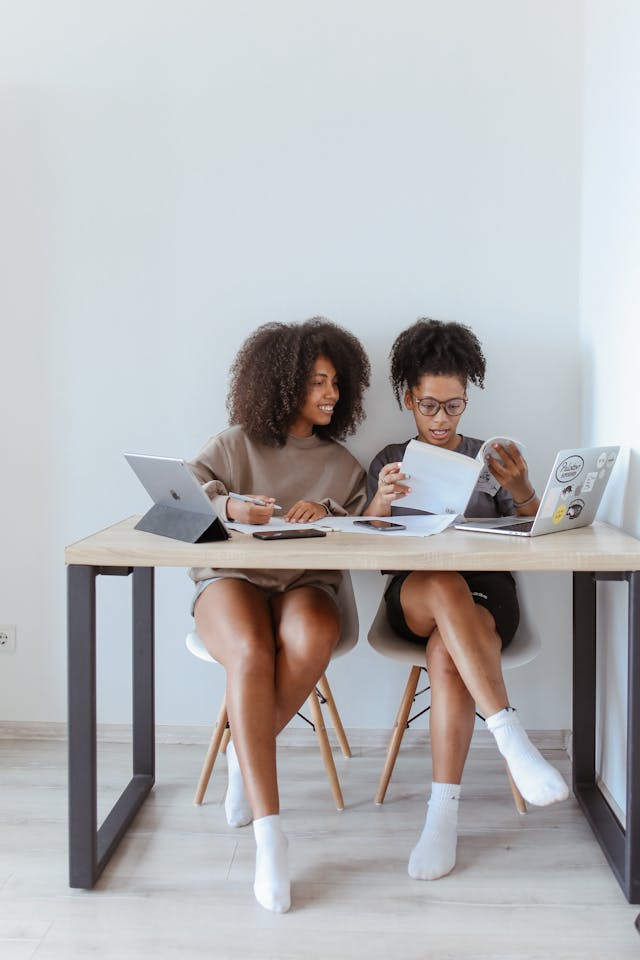 Dos mujeres están sentadas en un escritorio con sus computadoras portátiles.