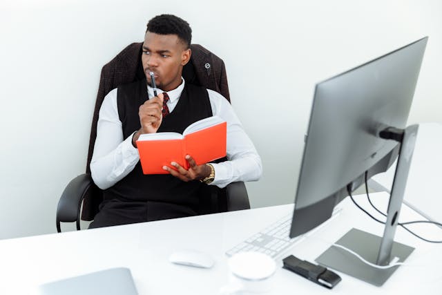 한 남성이 사무실에 앉아 컴퓨터 앞에 앉아 노트북과 펜을 들고 있습니다.
