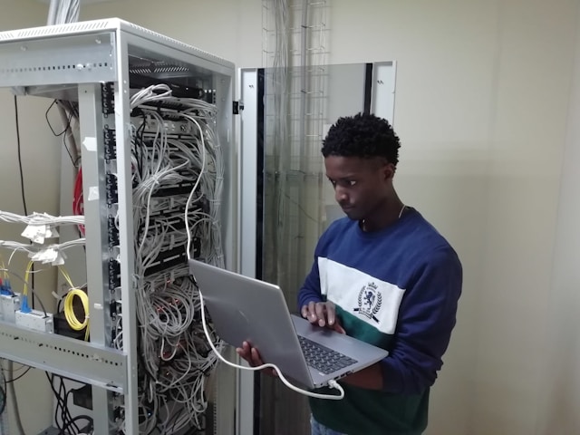 A person in a blue sweater checks a server with a laptop.
