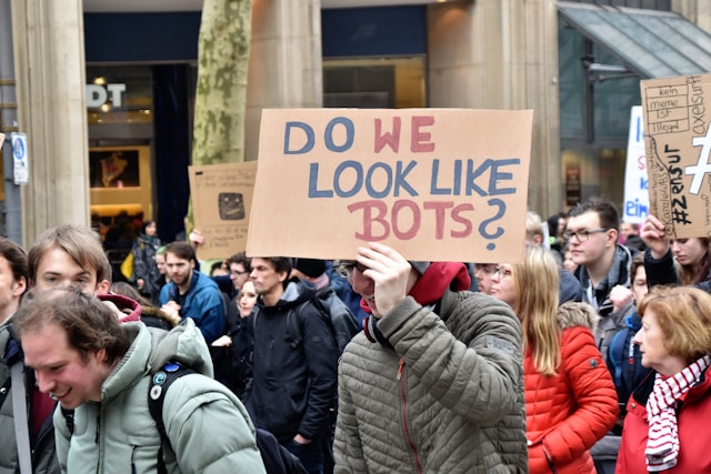 A person holds up a sign that says, “Do we look like bots?”
