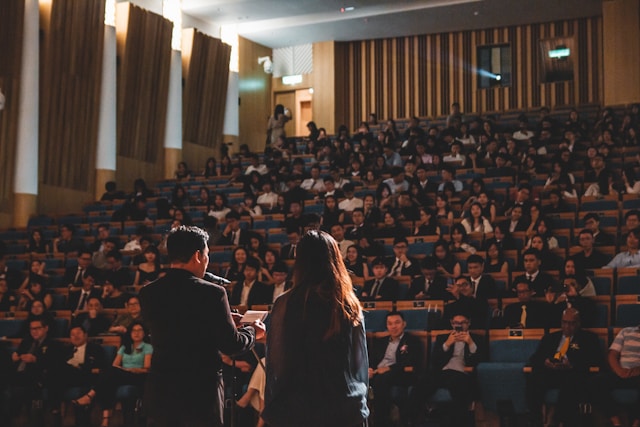 Duas pessoas falam diante de um grande público em uma sala de conferências. 