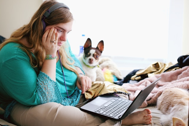 A woman with headphones sits on the bed with her dog and looks mystified as she scrolls through her TikTok page.