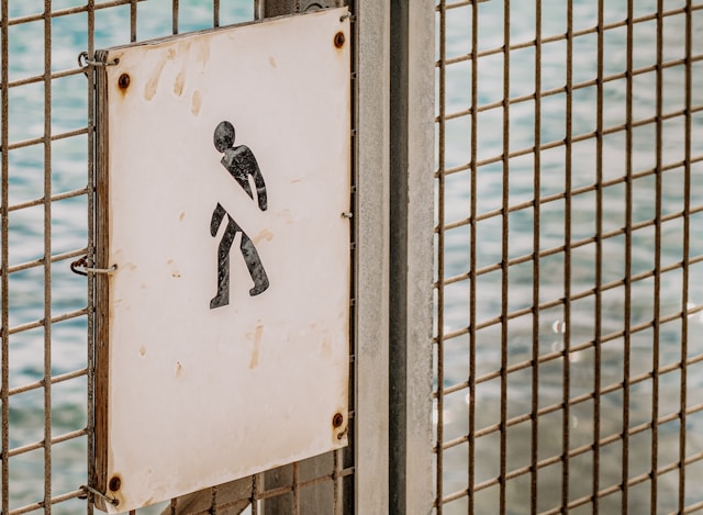 Un semn decolorat de pe o poartă metalică arată silueta unei persoane cu un simbol blocat deasupra ei.