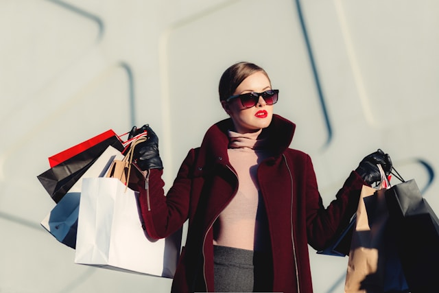 A fashion influencer in an expensive outfit carries shopping bags in both hands. 
