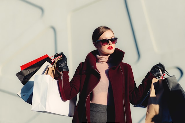 짙은 빨간색 코트와 선글라스를 쓴 여성이 양손으로 쇼핑백을 들고 있습니다. 