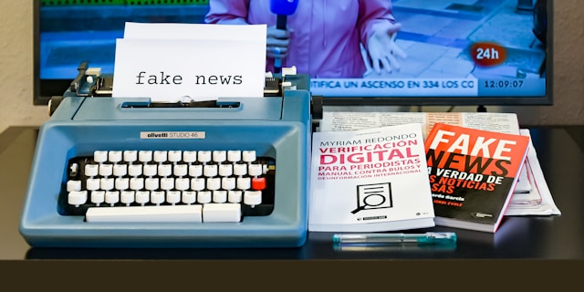Uma mesa está coberta com livros sobre notícias falsas e uma máquina de escrever com um pedaço de papel impresso com a inscrição "fake news".