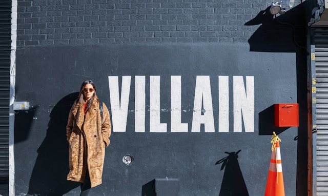 茶色の毛皮のコートを着た女性が、"Villain "と描かれた壁に向かって立っている。 