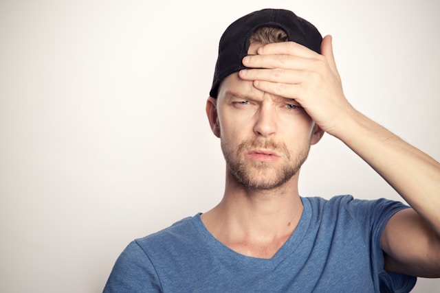 A man in a black cap and blue shirt puts a hand on his forehead and looks upset.
