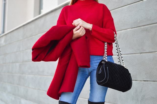 A fashion influencer wears a red sweater, jeans, and black knee-high boots while she carries a red jacket and black bag. 
