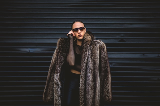 A woman wears an oversized dark brown fur coat over a sheer black top and jeans.